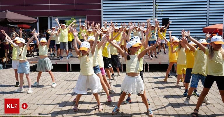 Eine Gruppe von Kindern in gelben Hemden und weißen Mützen führt einen Tanz vor einem Gebäude auf. Die Kinder heben ihre Hände und lächeln. Die Bühne hat einen roten Boden.