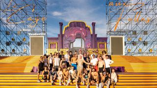 Eine Gruppe von Menschen posiert für ein Foto auf einer gelben Bühne mit einem Vaudeville Revue-Hintergrund. Einige tragen Hüte, Sonnenbrillen und Turnschuhe. Die Bühne hat ein rosa und lila Design.
