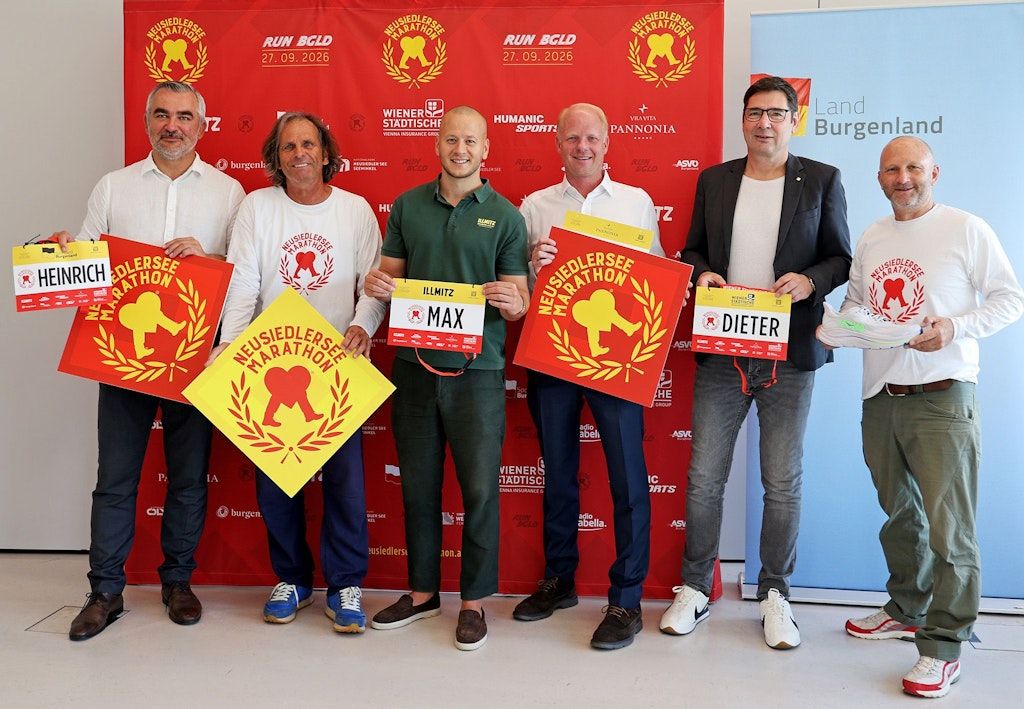 Fünf Männer posieren für ein Foto, halten Schilder und Zertifikate. Zwei halten Schilder mit dem Text 'Neusiedlersee Marathon' und einer Illustration eines rennenden Affen. Der Mann in der Mitte hält ein Zertifikat mit dem Namen 'Max'. Der Hintergrund hat rote Banner mit Logos und Text.