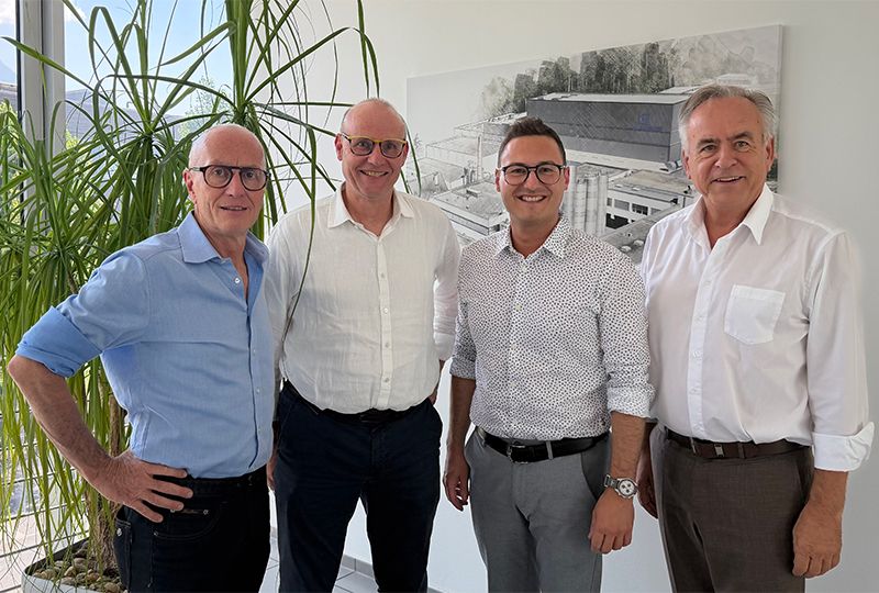 Vier Männer in formeller Kleidung, zwei mit Brille, stehen vor einem großen Bild. Einer trägt eine Uhr.