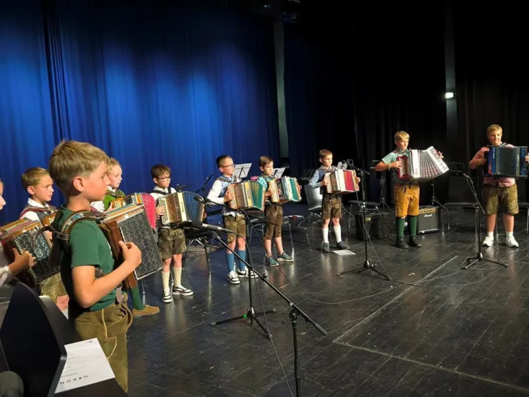 Eine Gruppe von Kindern spielt Akkordeons auf der Bühne. Der Junge vorne trägt Grün. Vier Jungen dahinter tragen traditionelle bayerische Kleidung.