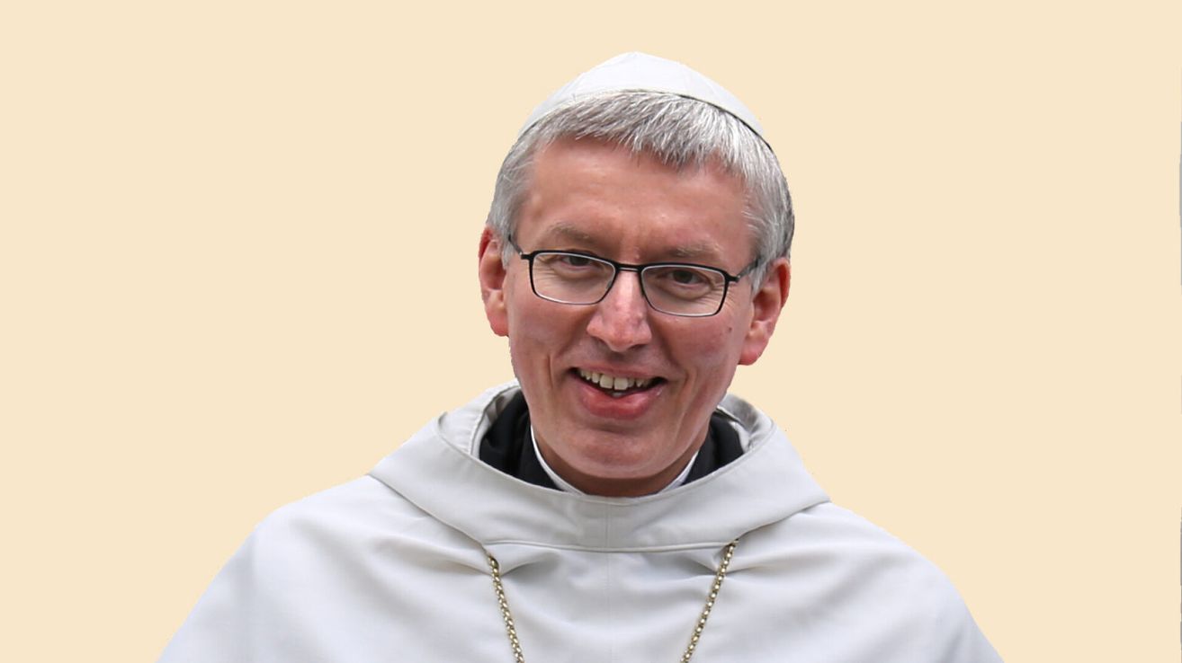 Ein Mann mit Brille, weißem Gewand und weißer Haube lächelt in die Kamera. Er hat eine Kette um den Hals.