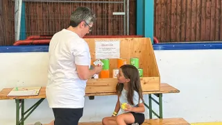 Eine ältere Frau steht neben einem jungen Mädchen an einem Tisch mit Dosen. Das Mädchen hält einen gelben Ball. Sie sind in einer Turnhalle.