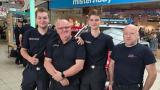 Vier Personen in schwarzen Uniformen stehen vor einem Mister Lady-Laden. Ein Polizeiauto ist dahinter geparkt.