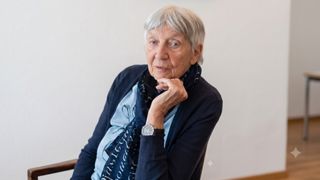 Eine ältere Frau mit grauem Haar, in einem blauen Pullover und einem Schal, sitzt mit der Hand am Kinn und sieht zur Seite. Sie trägt eine Uhr am linken Handgelenk.
