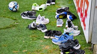 Eine Sammlung verschiedener Laufschuhe ist auf dem Rasen verstreut, darunter Nike, Puma und andere Marken. Einige Schuhe sind gepaart, andere nicht. Ein Fußball und ein Fußballhelm sind ebenfalls vorhanden.