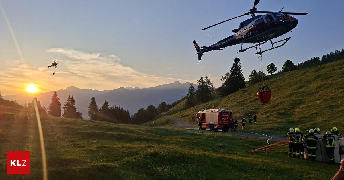 A police helicopter hovers above a fire truck on a mountain road, with a rescue basket hanging below. People are gathered around the truck.
