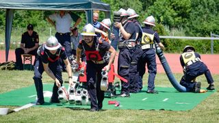 Eine Gruppe Feuerwehrleute in voller Uniform, die Feuerlöscher und Schläuche halten, führen eine Trainingsübung auf einem grünen Mattenbereich durch. Andere Feuerwehrleute beobachten von hinten.