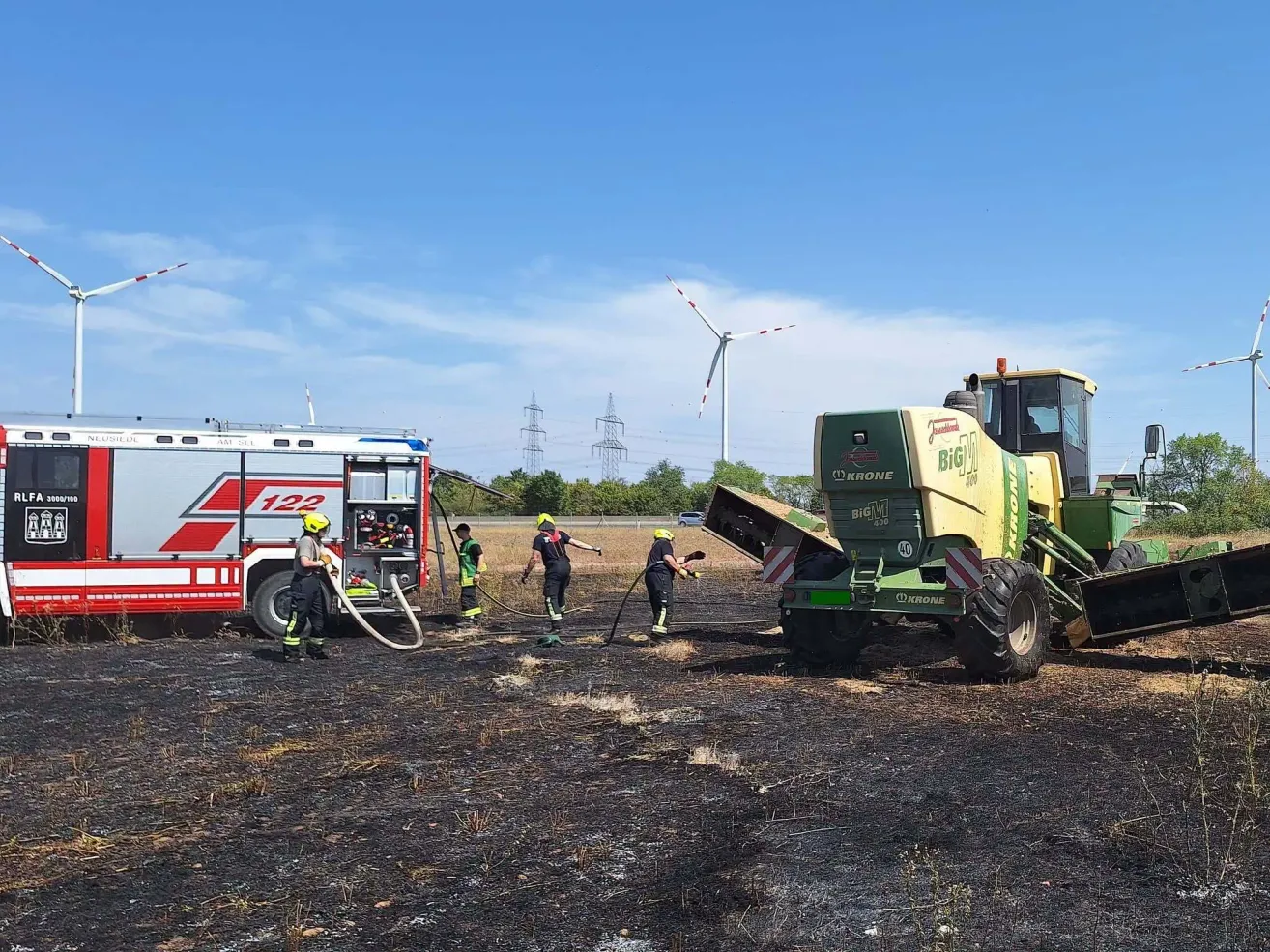 Feuerwehrleute löschen einen Brand an einem Mähdrescher mit einer Schlauchleitung, unter einem blauen Himmel mit einer Windturbine im Hintergrund.