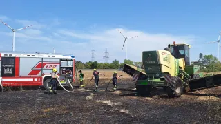 Feuerwehrleute löschen einen Brand an einem Mähdrescher mit einer Schlauchleitung, unter einem blauen Himmel mit einer Windturbine im Hintergrund.