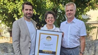 Drei Personen, zwei Männer und eine Frau, stehen zusammen mit einem gerahmten Zertifikat. Die Frau trägt eine Brille, und alle lächeln für die Kamera. Das Zertifikat ist für Grenzlandhof Reumann, datiert 2022.