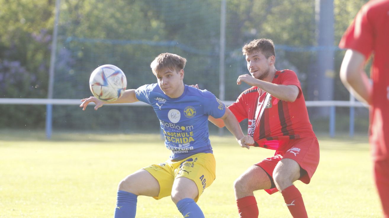 Zwei Fußballspieler in blauen und roten Trikots spielen auf einem Feld, einer versucht, den Ball zu blocken.