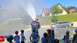 Ein Mann mit einem Feuerwehrschlauch steht auf der Straße und sprüht Wasser, das einen Regenbogen erzeugt. Kinder beobachten das Wasser von vorne. Dahinter steht ein Haus mit Solarpanelen.