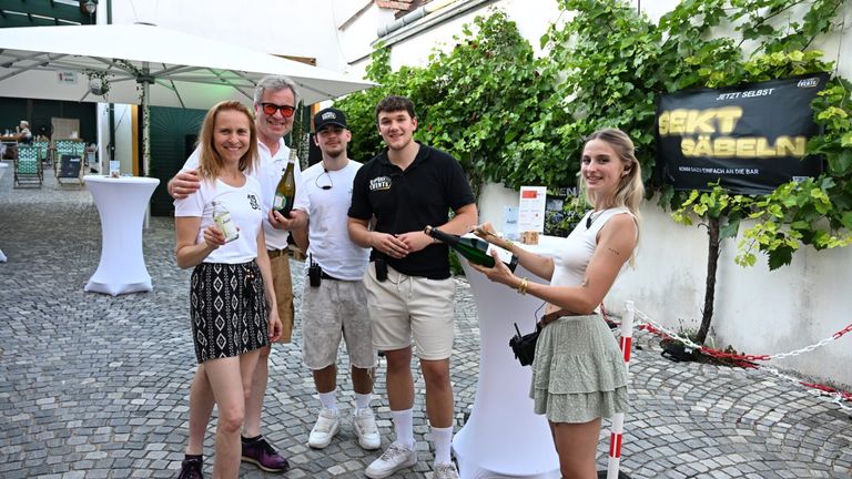 Fünf Personen posieren für ein Foto im Freien. Zwei Männer und zwei Frauen stehen vor einem weißen Tisch mit einer Flasche Wein darauf. Sie lächeln.