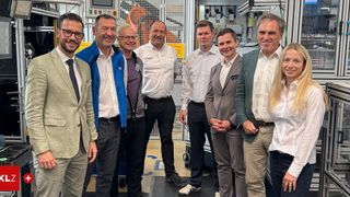 Eine Gruppe von Männern und Frauen, die formell gekleidet sind, lächeln und stehen zusammen vor einer Industriemaschine. Sie befinden sich in einer Fabrik.
