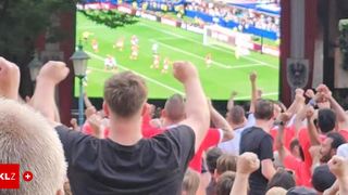 Eine Menge Fußballfans, einige mit erhobenen Armen, schauen ein Spiel auf einer großen Leinwand. Die Spieler sind auf dem Feld, und ein Tor ist sichtbar.