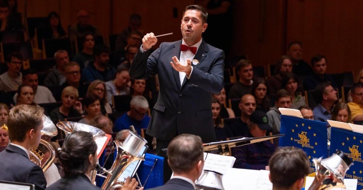 A man in a suit and bow tie conducts an orchestra in front of a crowd. The musicians play various instruments, including tubas and trumpets.
