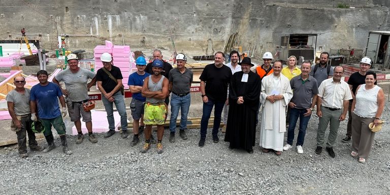 A group of people in hard hats and construction gear, including a priest, stand in front of a construction site.