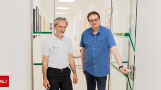 Zwei Männer, einer in einem weißen Poloshirt und der andere in einem blauen Hemd, stehen der Kamera gegenüber in einem Büroflur. Hinter ihnen befindet sich eine Glastür mit einem silbernen Griff.