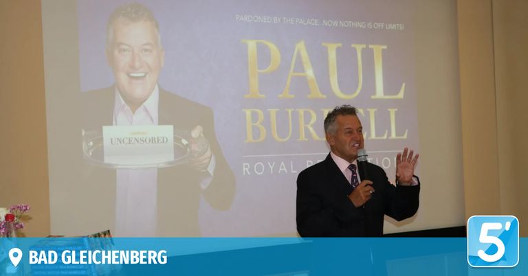 A man speaks into a microphone on stage in front of a large screen displaying the words 'Paul Burrell' and 'Uncensored'.