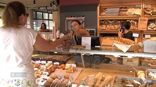 Eine Frau in einer Bäckerei bedient einen Kunden mit Gebäck. Hinter ihr steht eine Frau am Telefon. Regale voller Backwaren sind zu sehen.