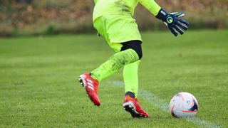 Ein Fußballspieler in einem neongrünen Trikot läuft mit Handschuhen auf den Ball zu. Der Ball ist weiß mit roten und schwarzen Markierungen.