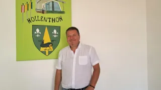 Ein Mann steht neben einem grünen Poster mit einem Haus und dem Namen Hollenthon darauf. Er trägt ein weißes Hemd und graue Hose.