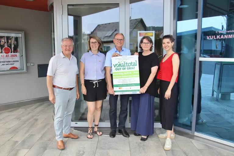 Five people pose outside a building with a sign that reads Vocal Total out of Graz. They are smiling and appear to be celebrating.