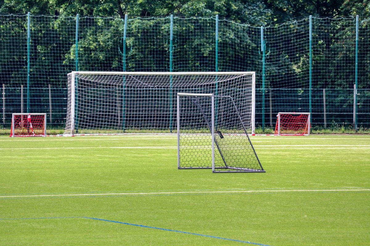 An empty soccer field with a white goal net and a red goal net on the right. Green fences surround the field. Trees are visible in the background.