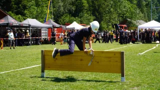 Eine Frau mit weißem Helm springt über eine Holzhurde auf einem Rasenfeld. Im Hintergrund befinden sich Menschen und Zelte.