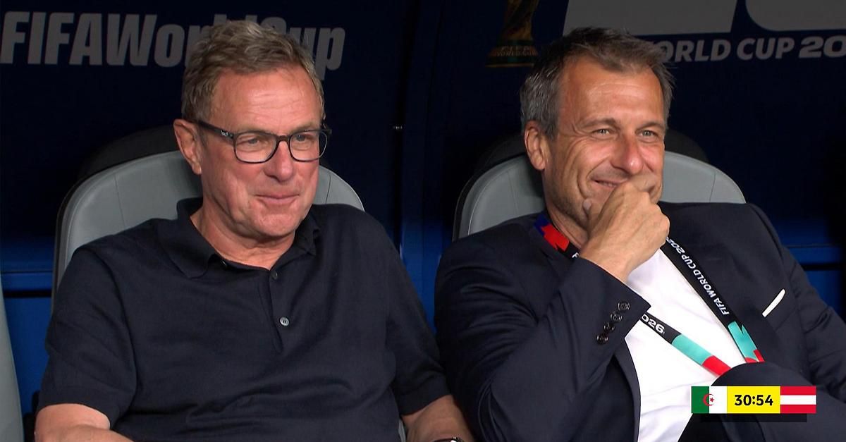 Zwei Männer sitzen in einem Stadion, einer trägt ein schwarzes Poloshirt, der andere einen Anzug mit weißem Hemd, beide lächeln und sehen in die Kamera.