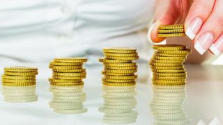 Eine Hand, die einen Stapel Goldmünzen über einem Tisch hält. Drei Stapel Münzen sind in aufsteigender Reihenfolge angeordnet.