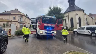 Feuerwehrleute in gelben Jacken gehen auf einen roten Feuerwehrwagen mit der Aufschrift 'FEUERWEHR' zu, dessen Türen offen sind. Die Szene spielt sich auf einer nassen Straße ab, mit einer Kirche im Hintergrund.