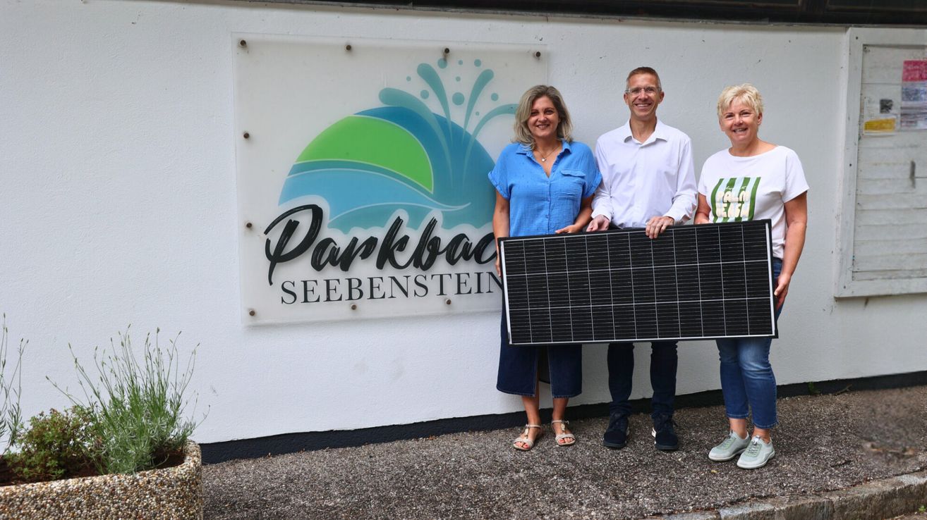 Drei Personen stehen vor einer Wand mit dem Schild Parkbad Seebenstein. Sie halten ein Solarpanel und scheinen zu lächeln.