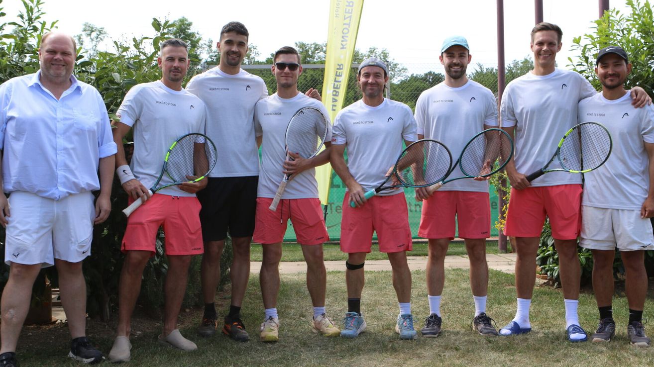 Fünf Männer in passenden weißen Shirts und korallenfarbenen Shorts posieren mit Tennisschlägern auf einer grasbewachsenen Freifläche. Der Mann ganz rechts trägt einen blauen Hut.
