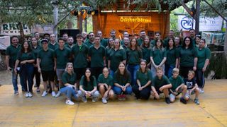 Eine Gruppe von Menschen in grünen Shirts und Hüten posiert für ein Foto auf einer Bühne mit einem hölzernen Hintergrund und Bäumen.