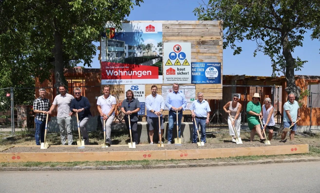 Eine Gruppe von Männern und Frauen hält Schaufeln und steht auf einer Erdstelle, möglicherweise während einer Grundsteinlegungszeremonie für ein Bauprojekt. Dahinter befindet sich ein großes Schild mit Logo und Text sowie ein Holzschild.
