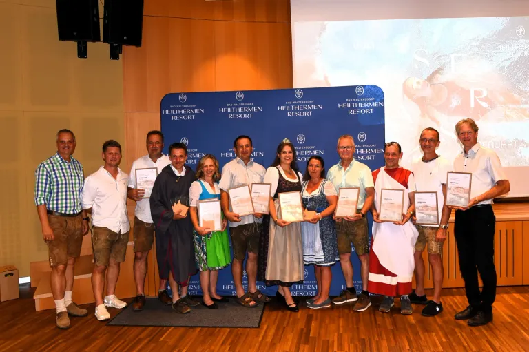 A group of people dressed in traditional clothing are posing for a photo in front of a blue backdrop with 'Bad Waltersdorf Heilthermen Resort' written on it.