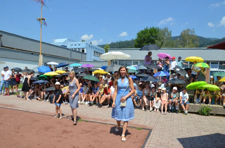 Zwei Frauen gehen vor einer Menschenmenge, viele mit bunten Schirmen. Im Hintergrund steht ein Schulgebäude unter blauem Himmel.