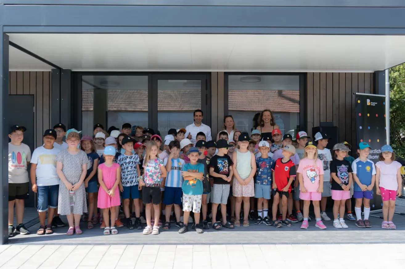 Eine Gruppe von Kindern und Erwachsenen steht vor einem modernen Gebäude. Die Kinder tragen Mützen, und Erwachsene stehen hinter ihnen. Der Mann in der Mitte lächelt für die Kamera.
