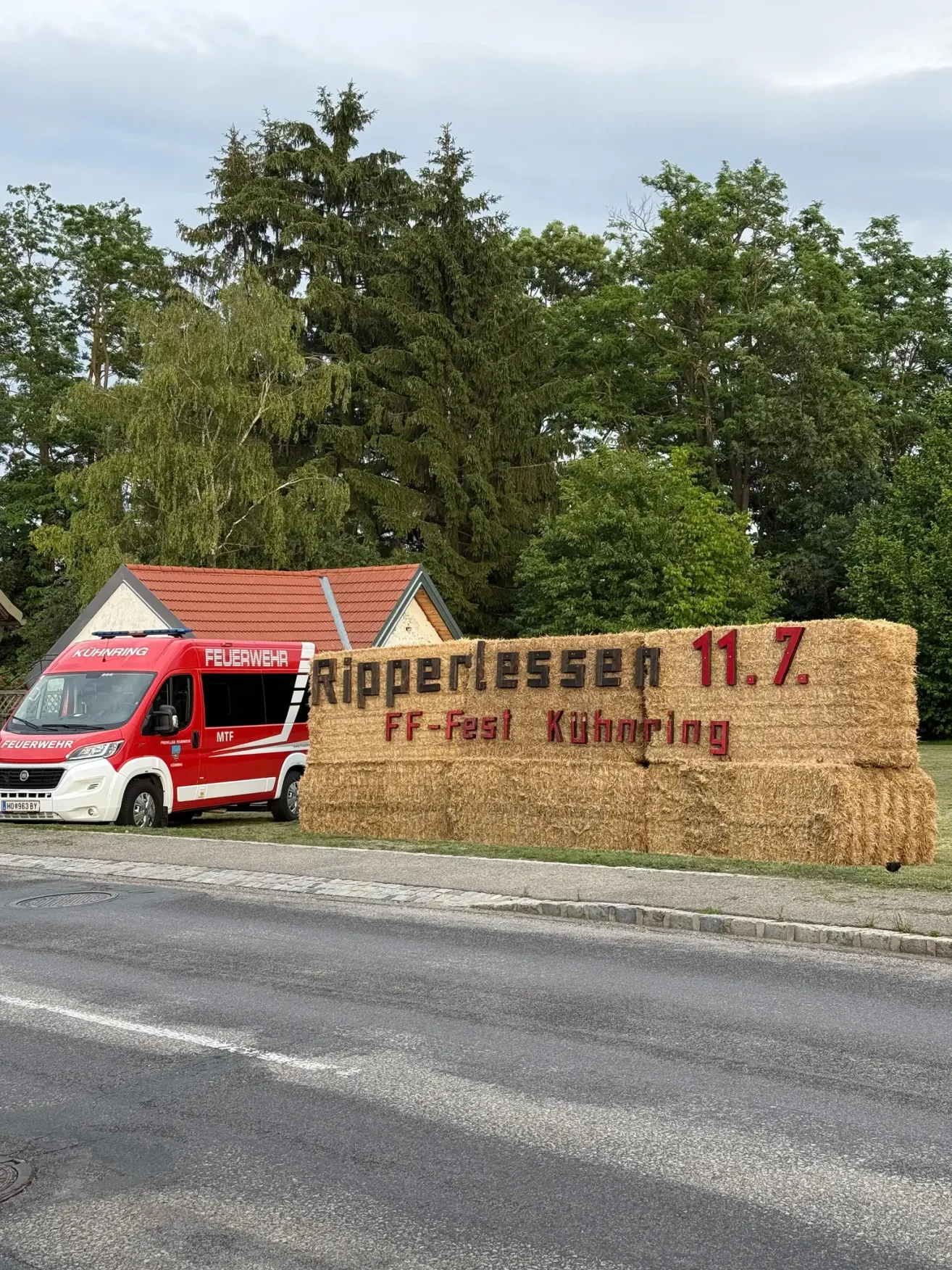 A red fire truck is parked next to a straw bale sign advertising the FF-Fest Kühnring in Ripperl. The sign reads 'Ripperlessen 11.7.' It's a sunny day with green trees in the background.