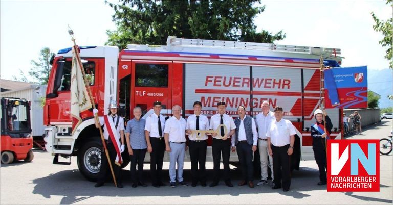 Several firefighters stand in front of a red fire truck, holding a key and posing for a photo. The truck has a sign that reads 'Feuerwehr Meiﬁningen'.