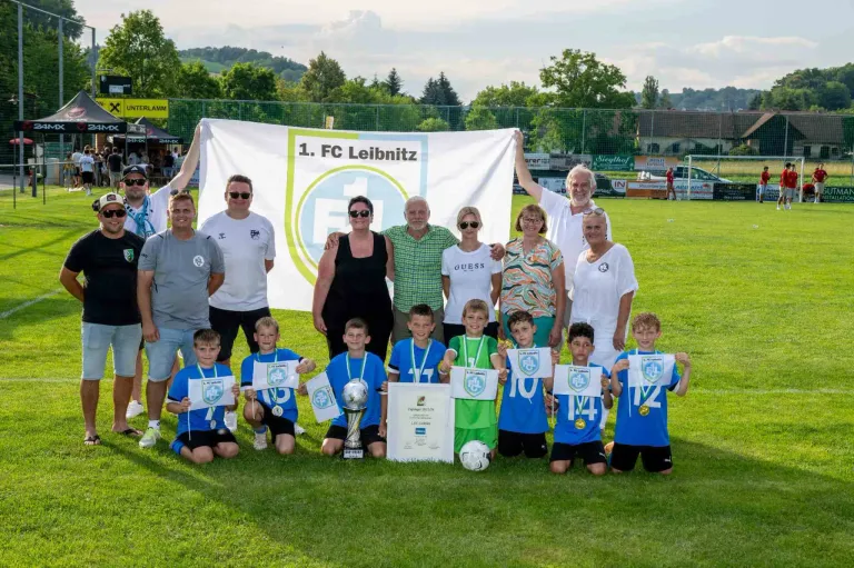 A soccer team with their coaches and parents pose for a photo with trophies and medals on a grassy field. A banner with 1. FC Leibnitz is held in the background.