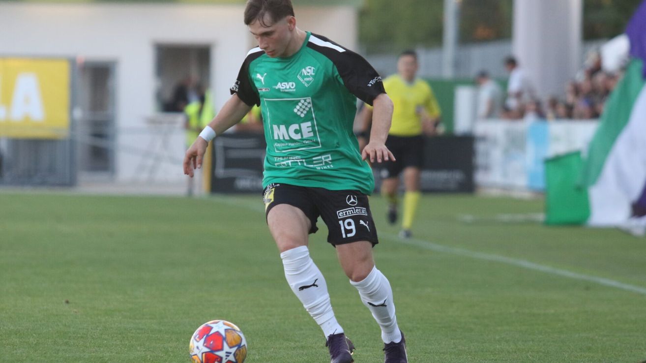 Ein Fußballspieler in einem grünen Trikot mit der Nummer 19 auf dem Rücken tritt einen Ball. Im Hintergrund sind andere Spieler und Personen verschwommen.