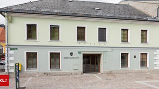 Ein weißes Gebäude mit einem grauen Dach, einem großen Eingang, einem Schild mit der Aufschrift 'BEZIRKSGERICHT' und mehreren Fenstern mit Jalousien.