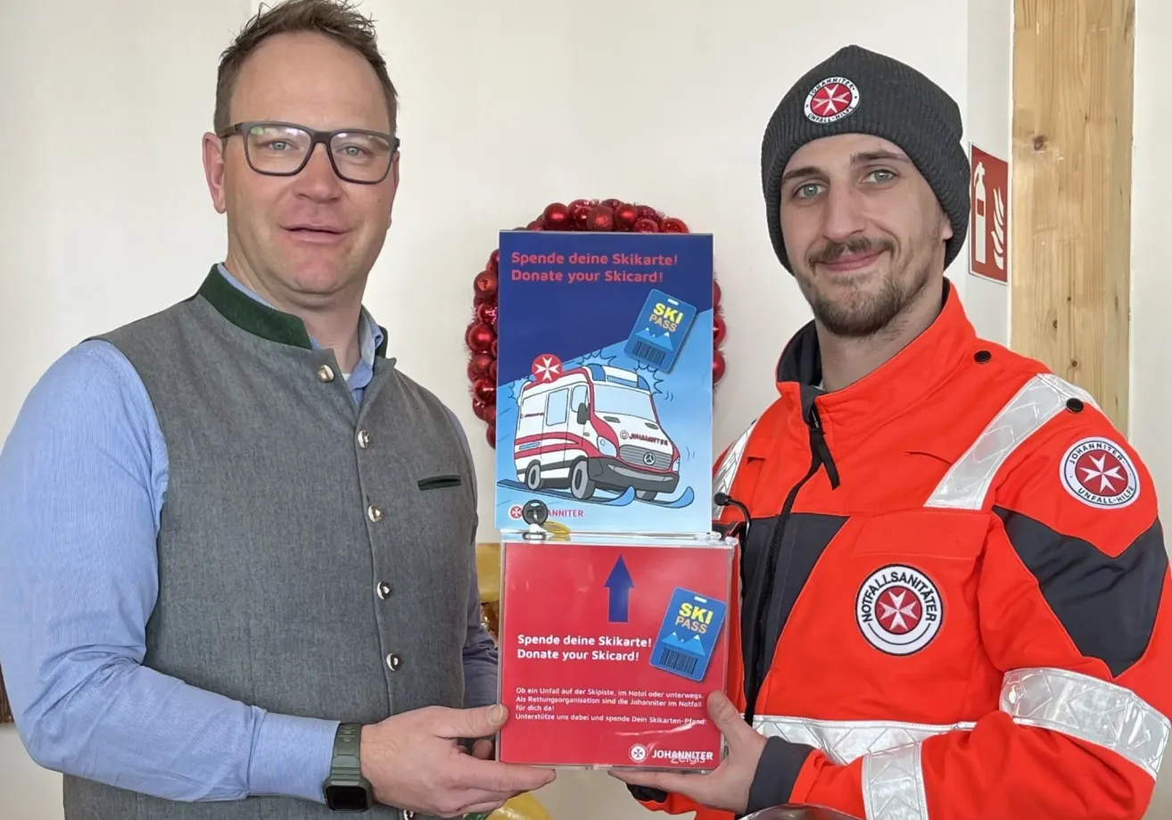 Two men hold donation boxes for ski passes. One wears glasses and a gray jacket, the other a beanie and an orange jacket.
