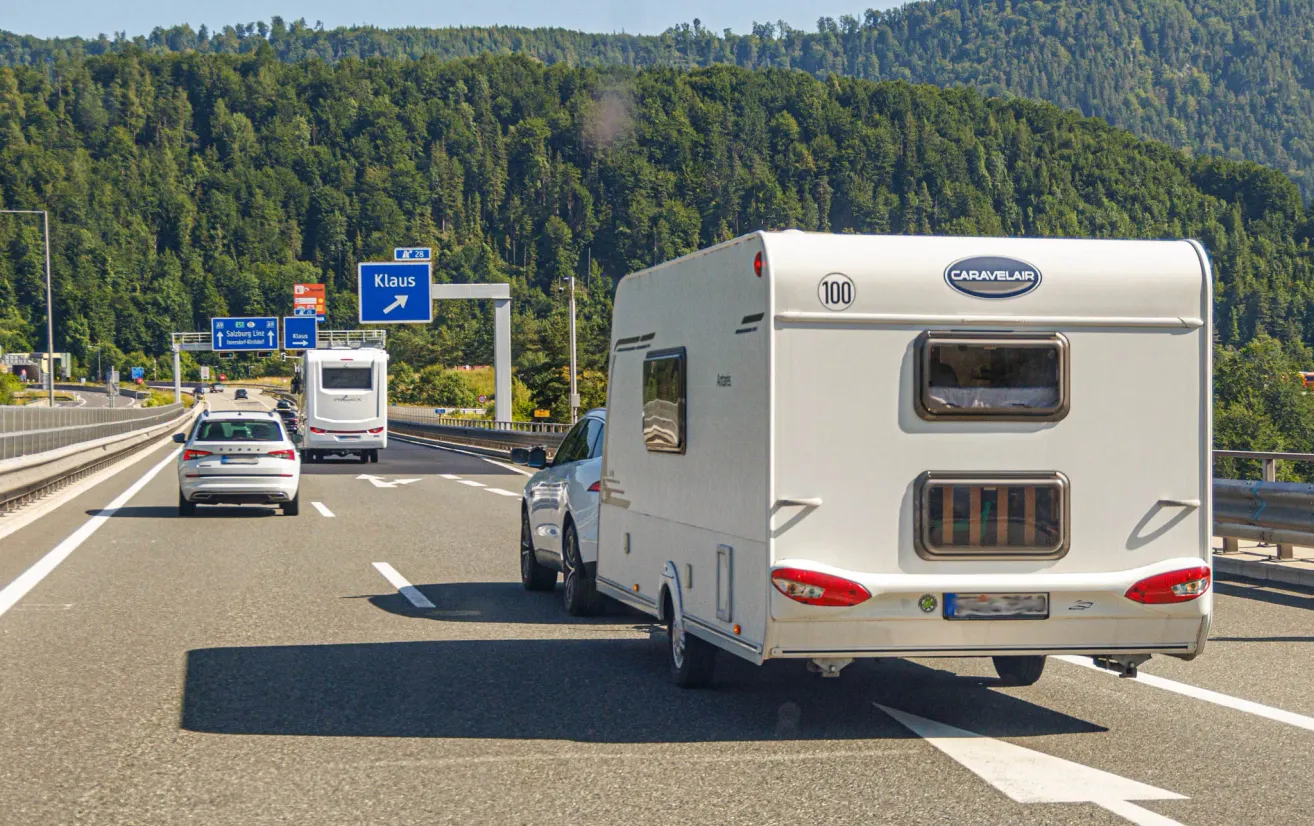 Ein weißer Caravelair-Anhänger fährt auf der Autobahn mit anderen Fahrzeugen. Im Hintergrund befinden sich Hügel mit einem Schild, das Klaus sagt.