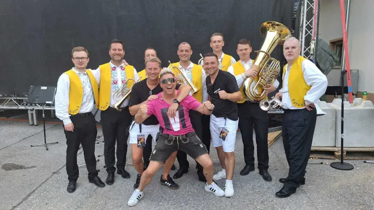 A group of musicians in yellow vests and black pants posing for a photo. They are holding musical instruments, including trumpets and tubas.