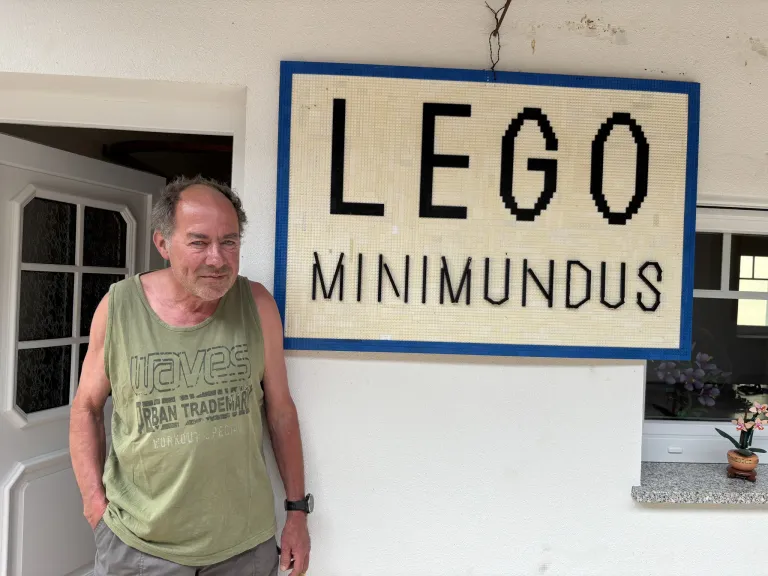 A man stands by a sign that reads LEGO MINIMUNUS, made from small square tiles. He wears a green tank top with waves and urban trademark.
