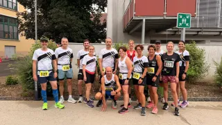 Eine Gruppe von Läufern posiert für ein Foto vor einem Gebäude, mit nummerierten Shirts und Turnschuhen, mit einem Balkon und einem grünen Schild im Hintergrund.